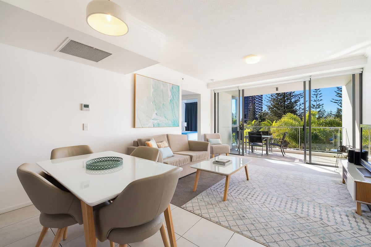 Una sala de estar moderna con comedor, ventanas amplias y vistas a árboles y edificios.