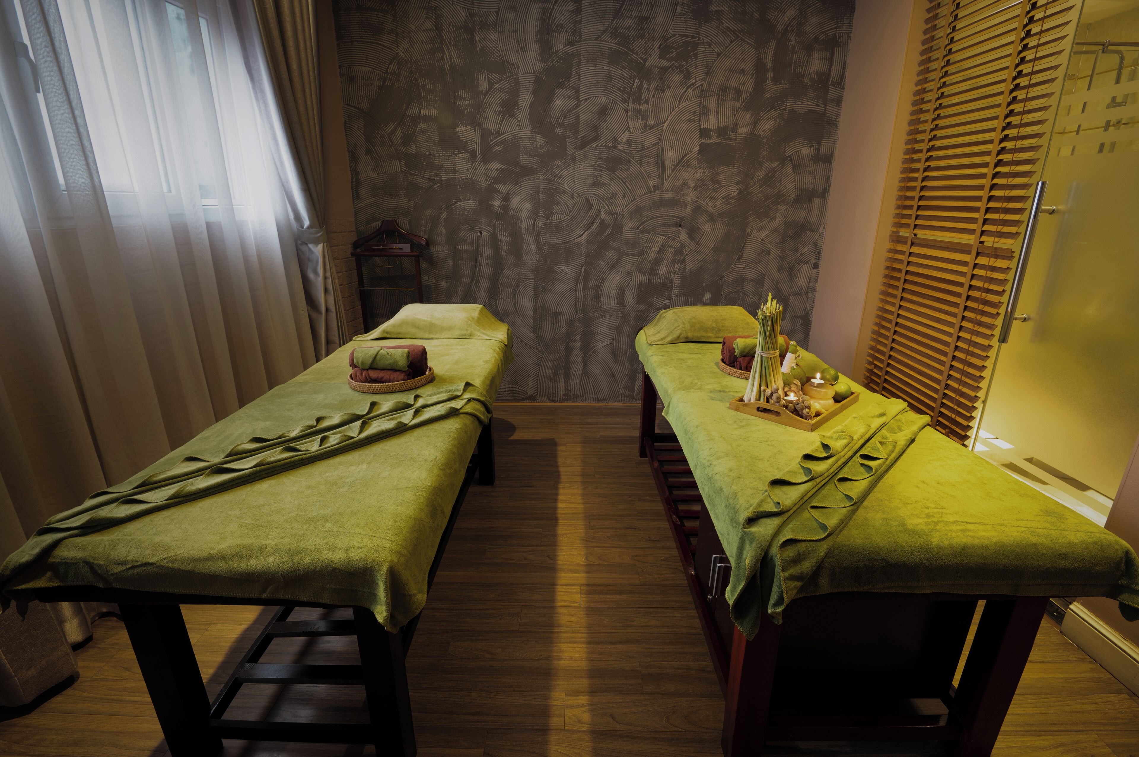 A room with two beds, each with a green bedspread and pillows, a wooden floor, and a patterned wall.