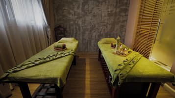 A room with two beds, each with a green bedspread and pillows, a wooden floor, and a patterned wall.