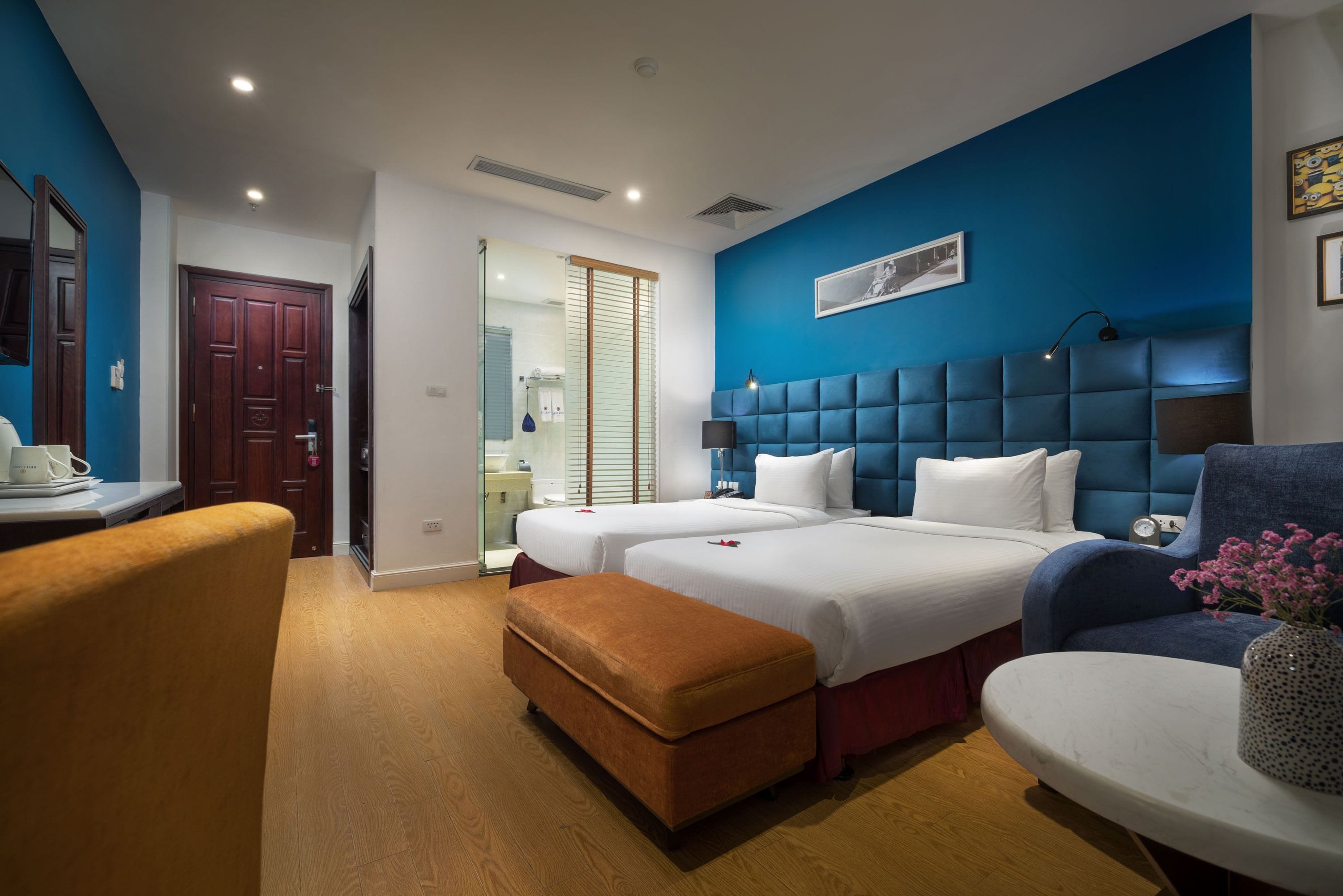 Ein Hotelzimmer mit zwei Betten, einem Schreibtisch, einem Stuhl und einem durch eine Glastür einsehbaren Badezimmer.