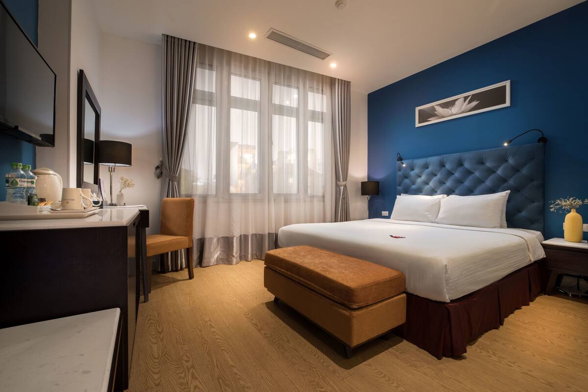 A hotel room with a large bed, a desk with a chair, a small table, and a window with curtains.