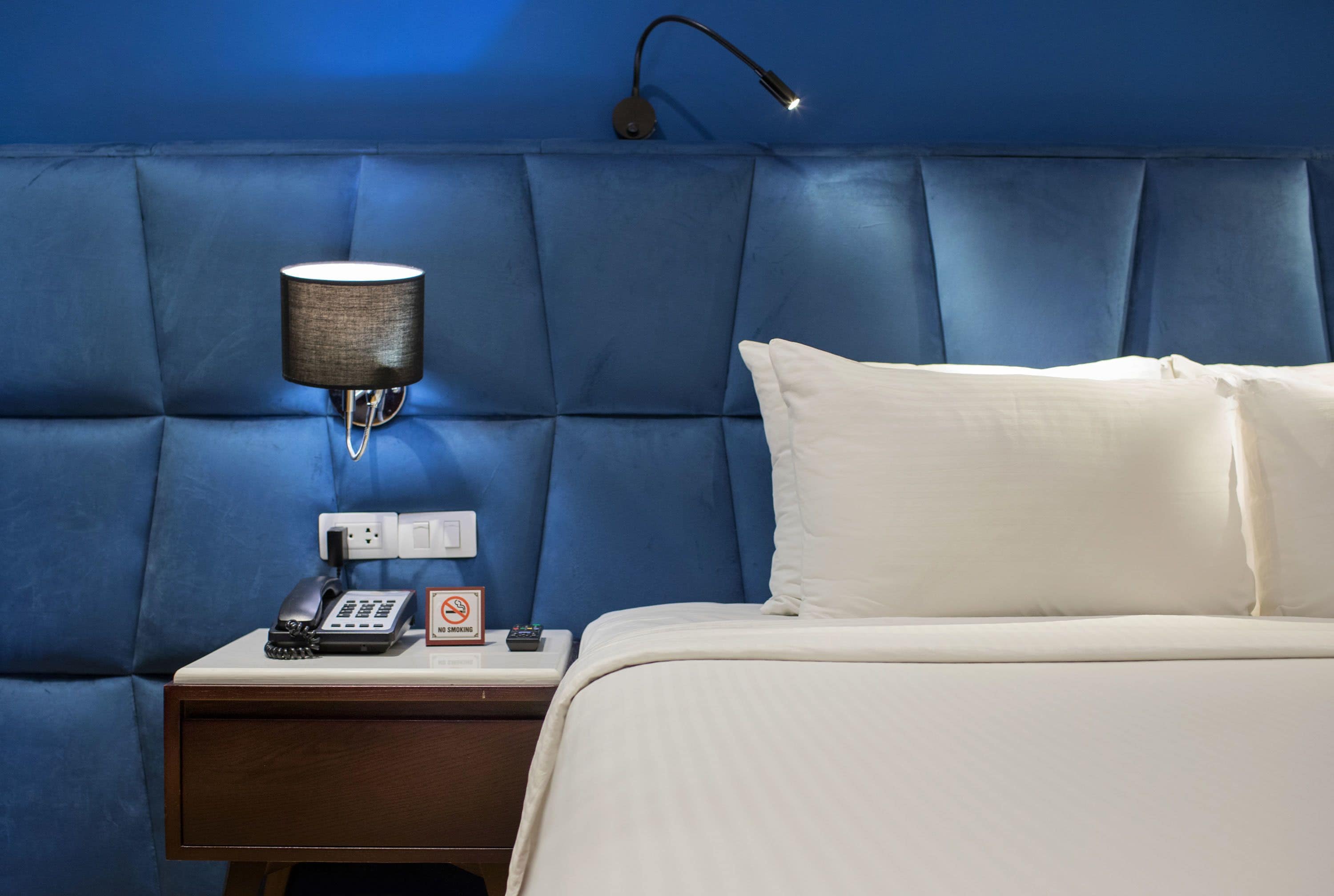 A hotel room with a blue padded headboard, a bedside lamp, a phone, a "No Smoking" sign, and a neatly made bed with white linens.
