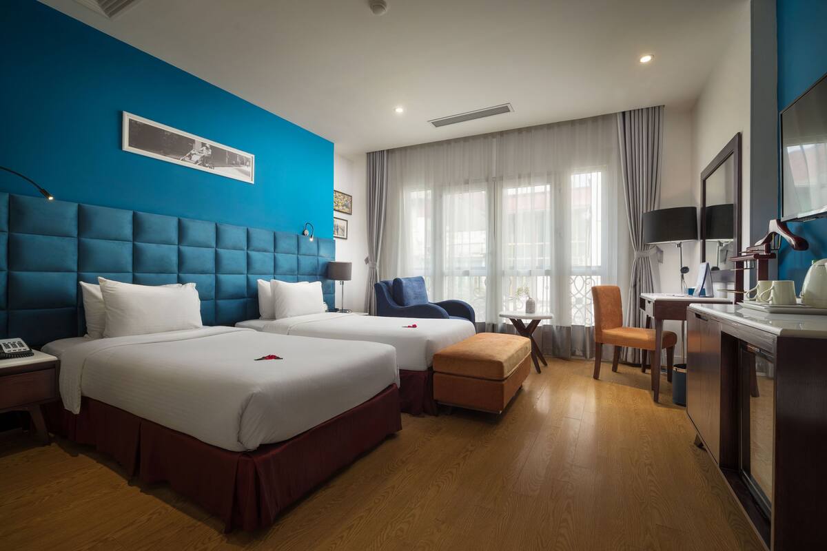 A hotel room with two beds, a desk, a chair, a small table, and a large window with curtains.