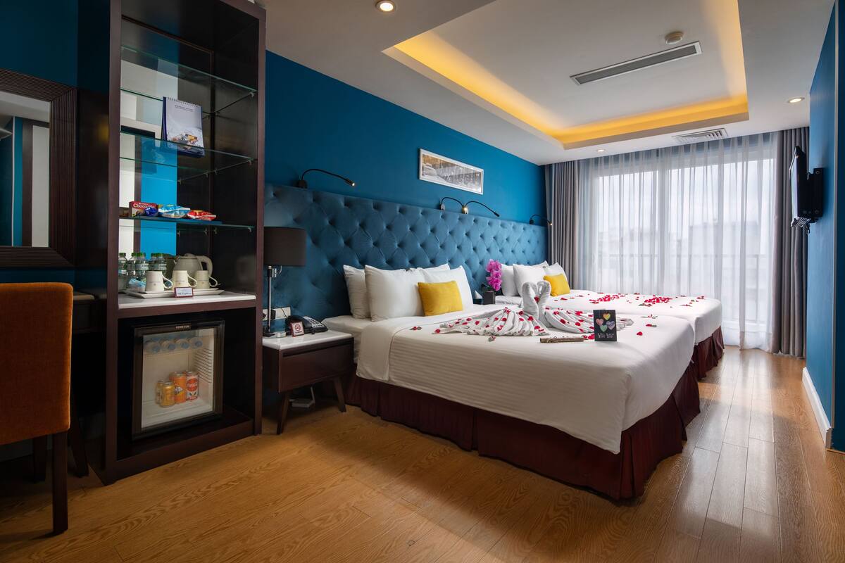 A hotel room with a large bed, a minibar, a desk with a lamp, and a large window with curtains.