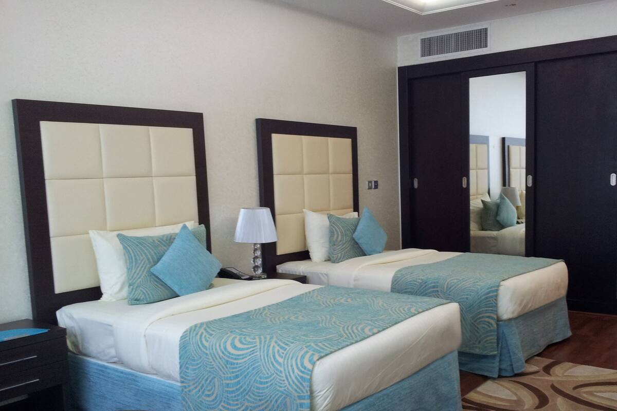 A hotel room with two beds, each with a headboard, a bedside table with a lamp, and a small table with a chair.