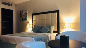A hotel room with a large bed, two bedside lamps, a small round table, a chair, and a painting on the wall. - Gokulam Park Doha (Doha)