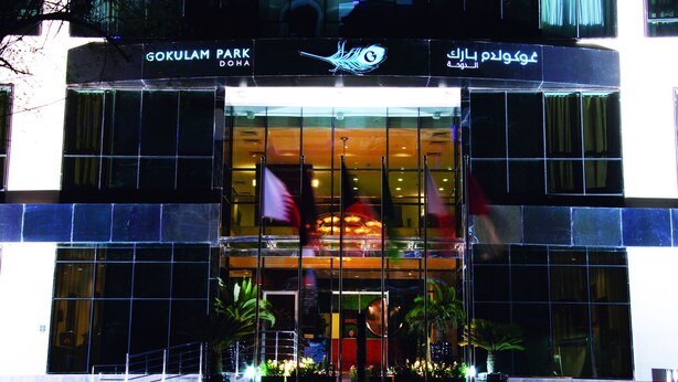 The exterior of GOKULAM PARK DOHA with a glass facade, potted plants, and flags. - Gokulam Park Doha (Doha)