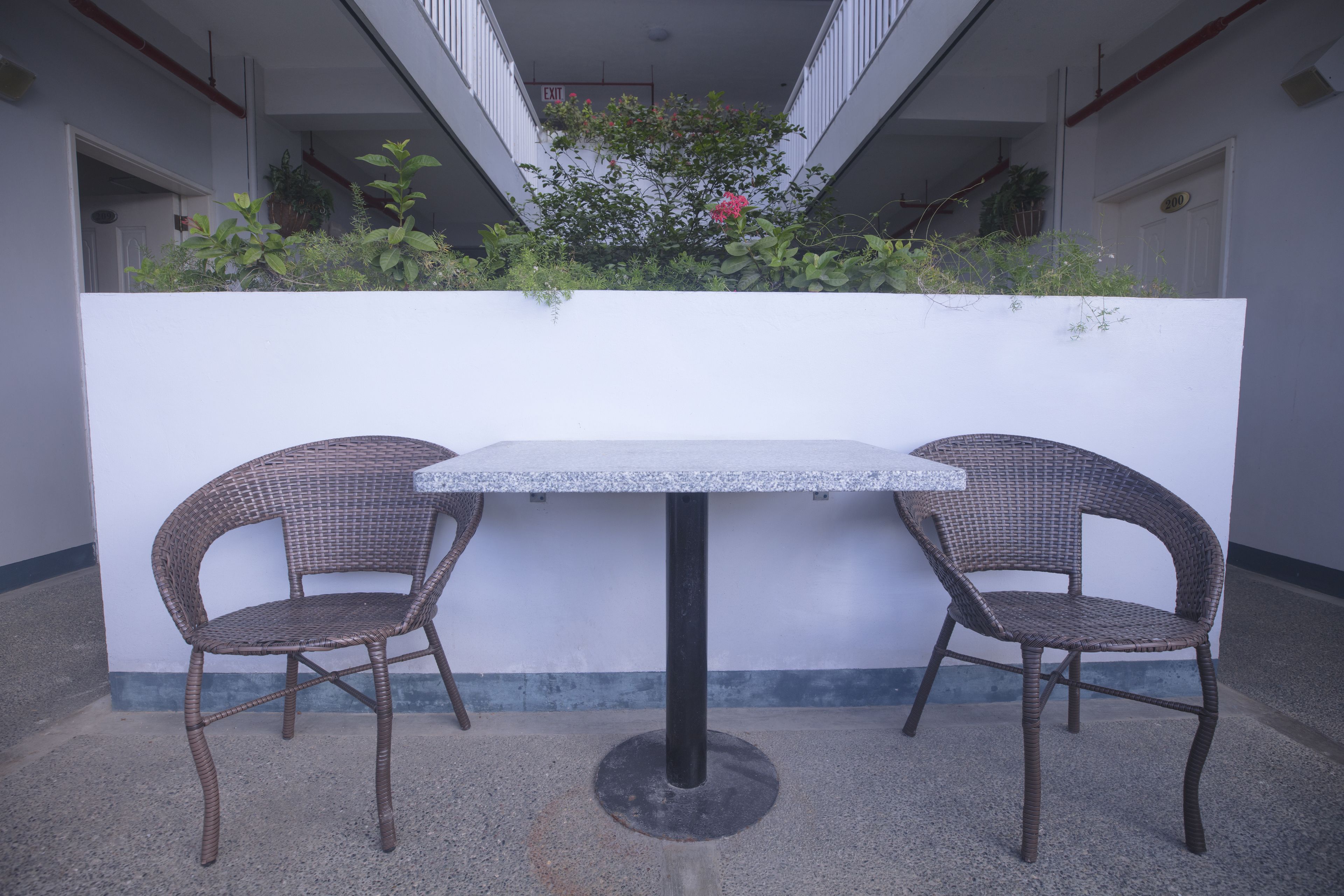 Un pequeño espacio al aire libre con una mesa y dos sillas de mimbre, rodeado por un muro blanco y un balcón con plantas.