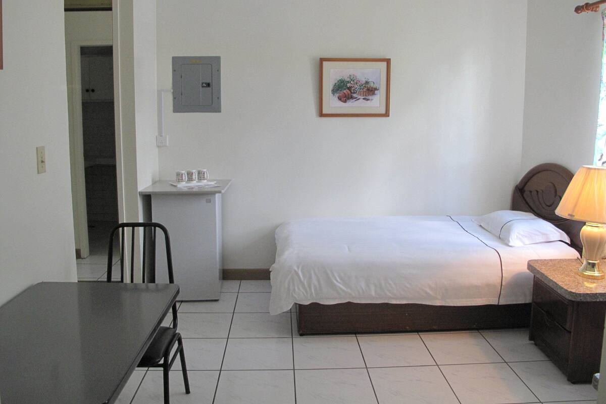 Una habitación pequeña y limpia con cama, escritorio, silla y mesita de noche con lámpara.