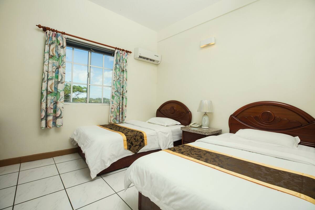Una habitación de hotel con dos camas, una ventana con cortinas, un equipo de aire acondicionado y una mesita de noche con lámpara.