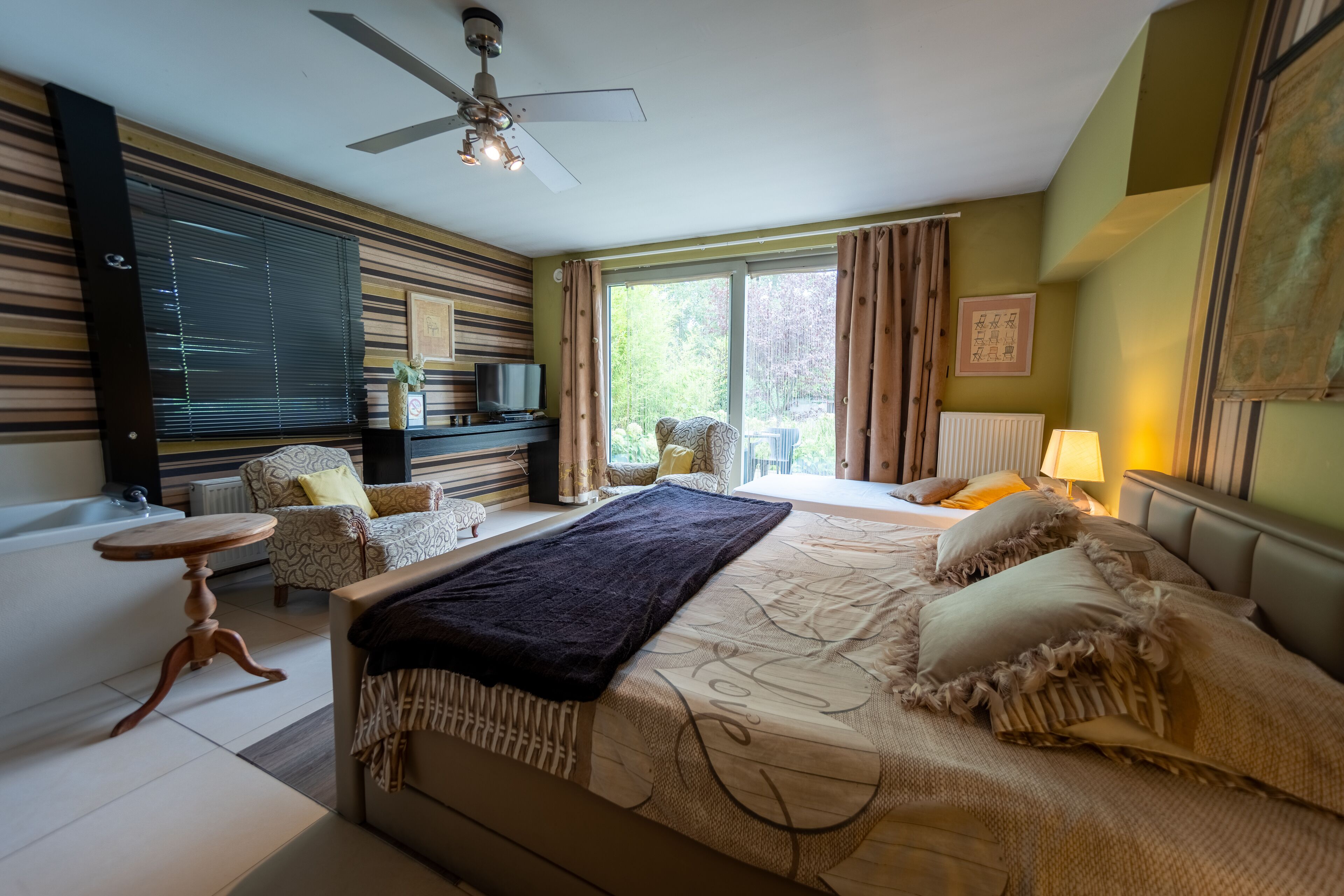 Un dormitorio con una cama grande, un ventilador de techo, una ventana con cortinas, un sofá y una mesita.