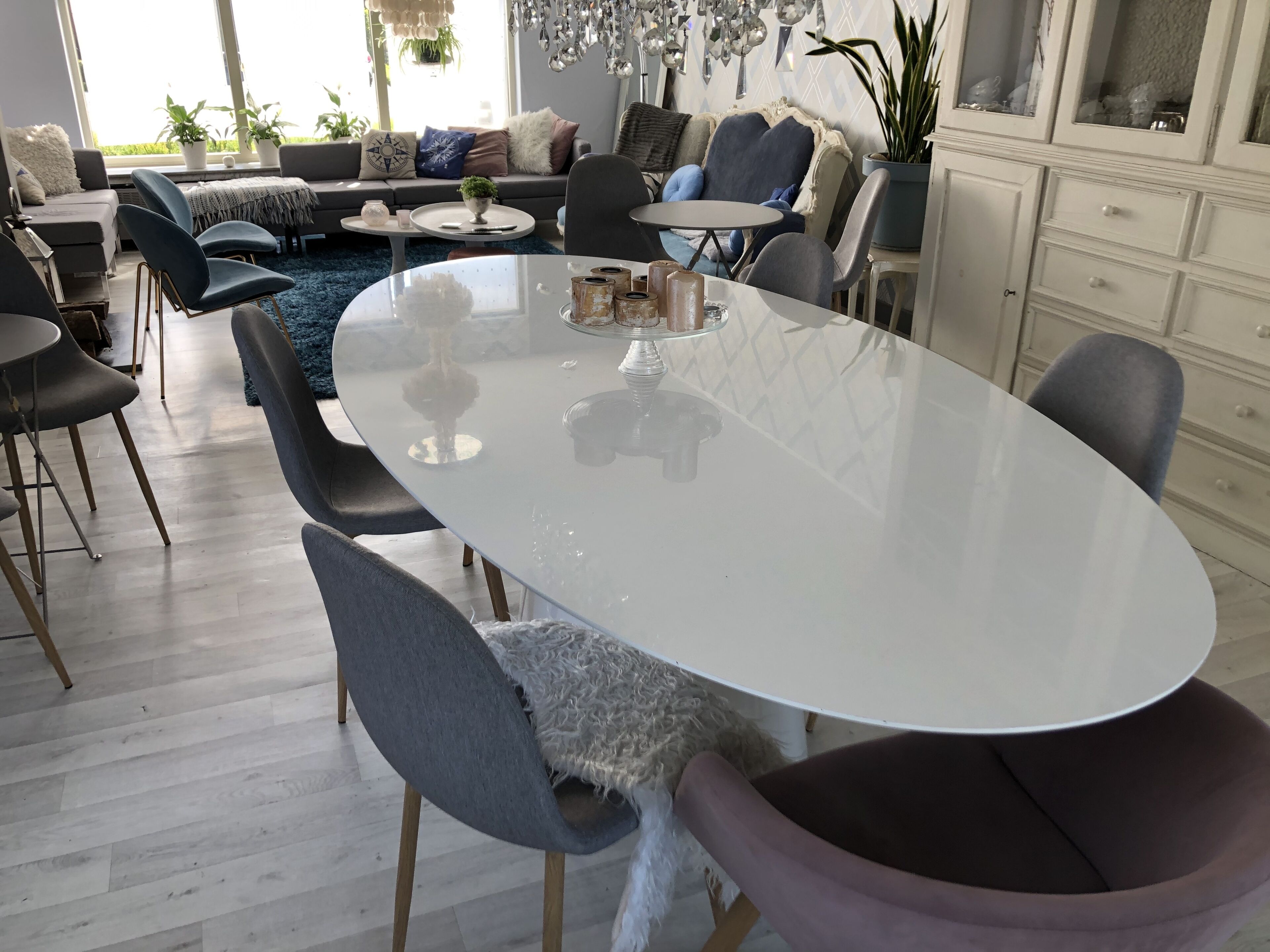 En moderne stue med et stort ovalt spisebord, grå polstrede stole, en sofa med mange puder, og et stort vindue der lukker naturligt lys ind.