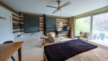 A bedroom with a bed, a desk, a chair, a TV, and a balcony with a view of greenery.