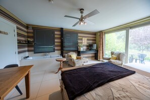 Un dormitorio con cama, escritorio, silla, televisor y balcón con vista a follaje.
