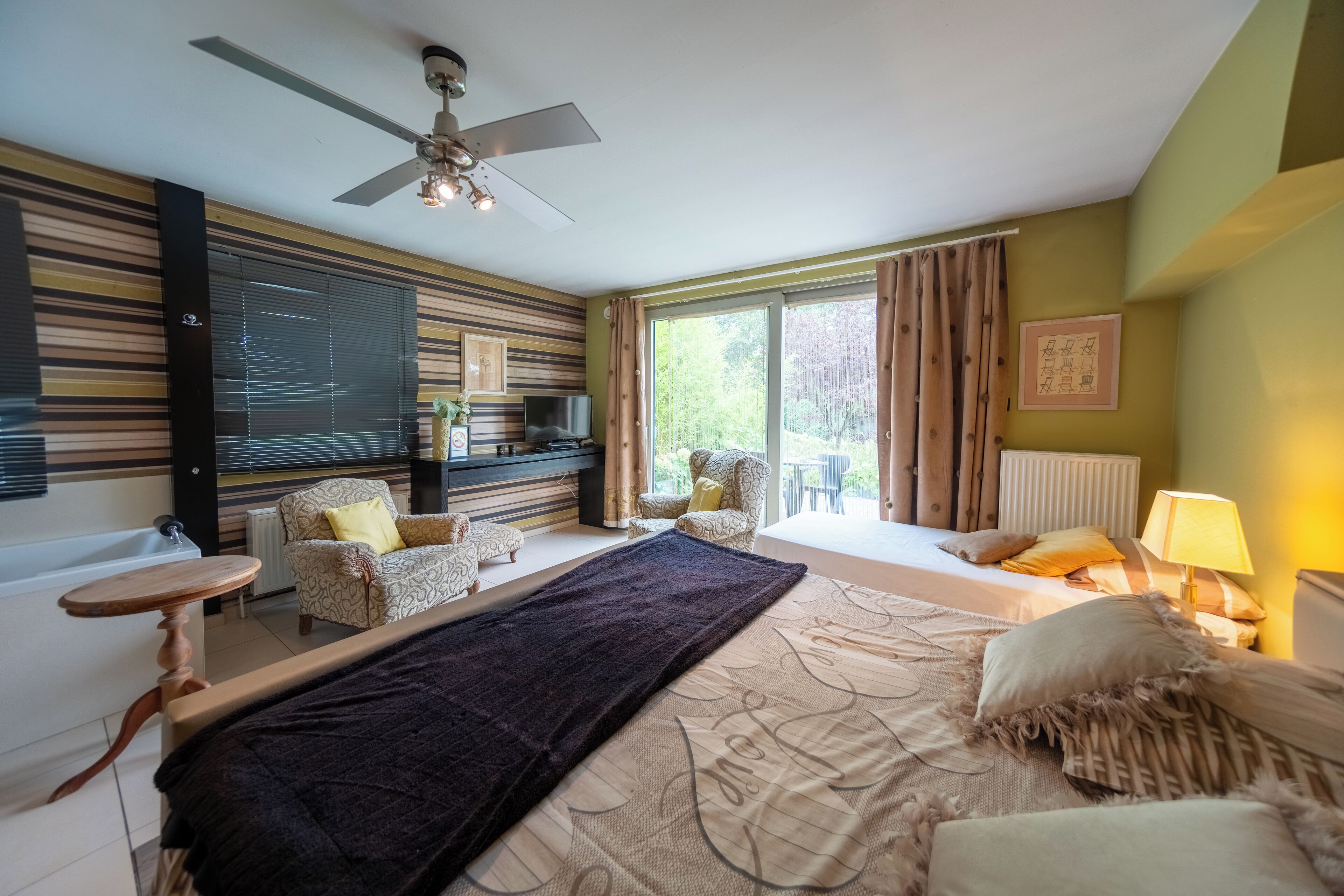 A bedroom with a large bed, a ceiling fan, a TV, a sofa, and a window with curtains.