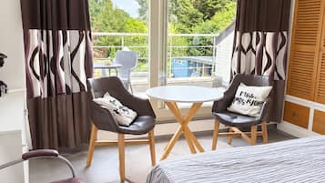 A hotel room with a bed, two chairs, a small table, a balcony with a view of greenery, and a modern chandelier.
