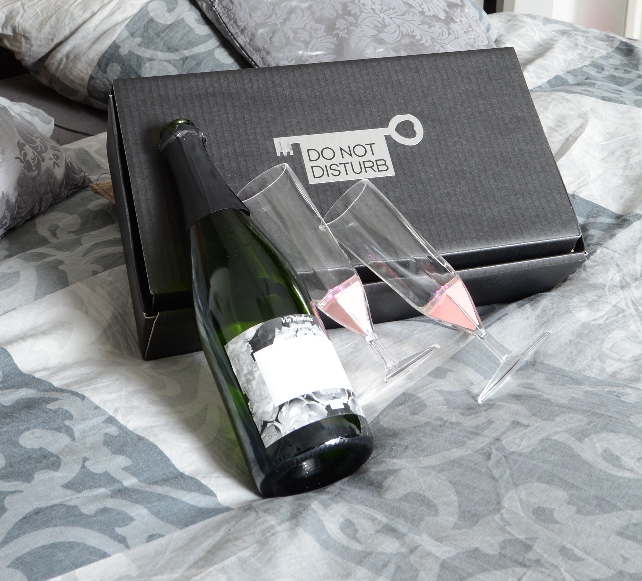 A bottle of wine, two glasses, and a black box with a label on a bed with patterned bedding.