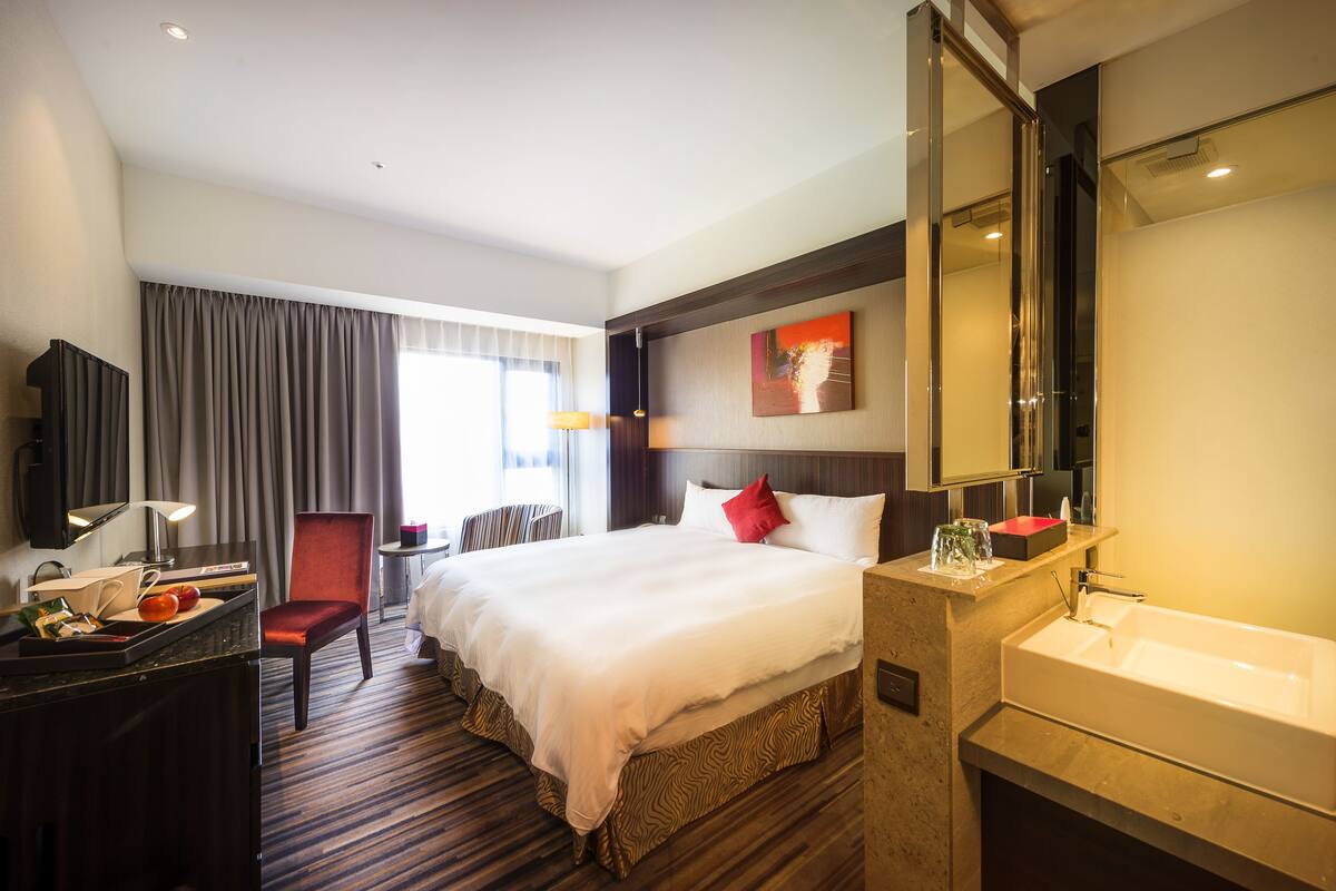 Habitación de hotel con una cama grande, una zona de estar con una silla, un escritorio con lámpara, un televisor y un baño con lavamanos y espejo.