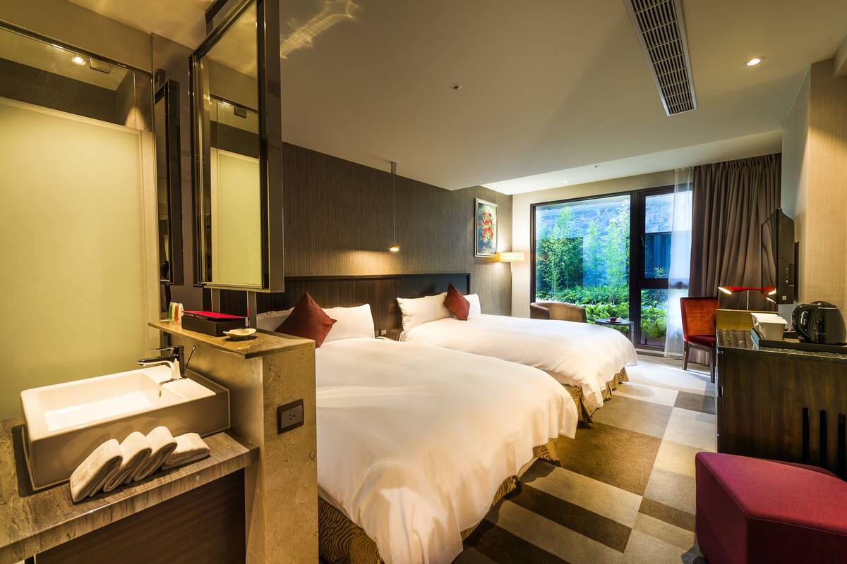 Habitación de hotel con dos camas, un ventanal grande, un escritorio y un baño con lavamanos y espejo.