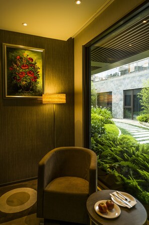 A modern room with a framed painting, a comfortable armchair, a small table with a plate of apples and cutlery, and a view of a landscaped garden through a large window.