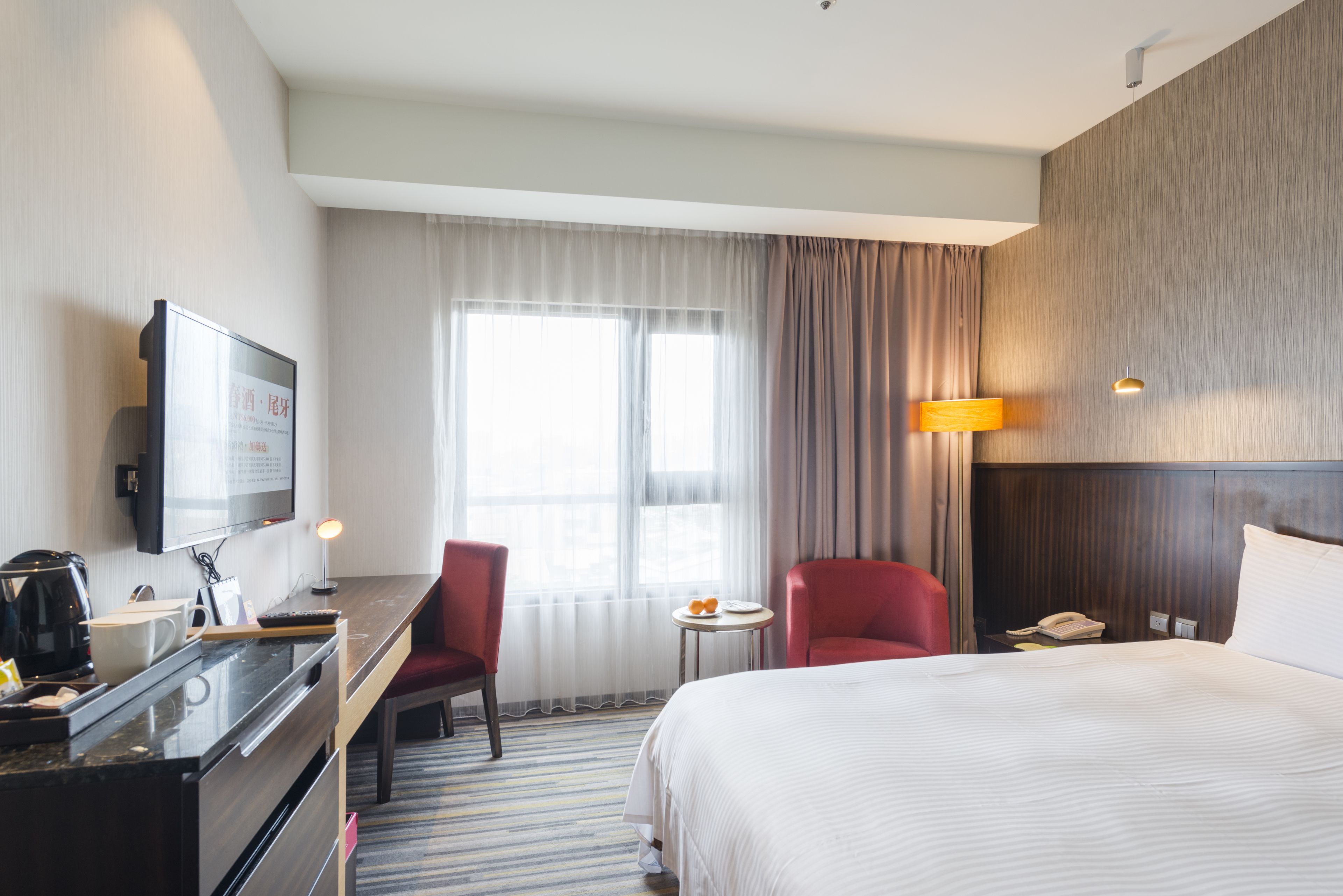 A hotel room with a bed, a desk with a computer, a TV, a chair, a small table with fruit, and a window with curtains.