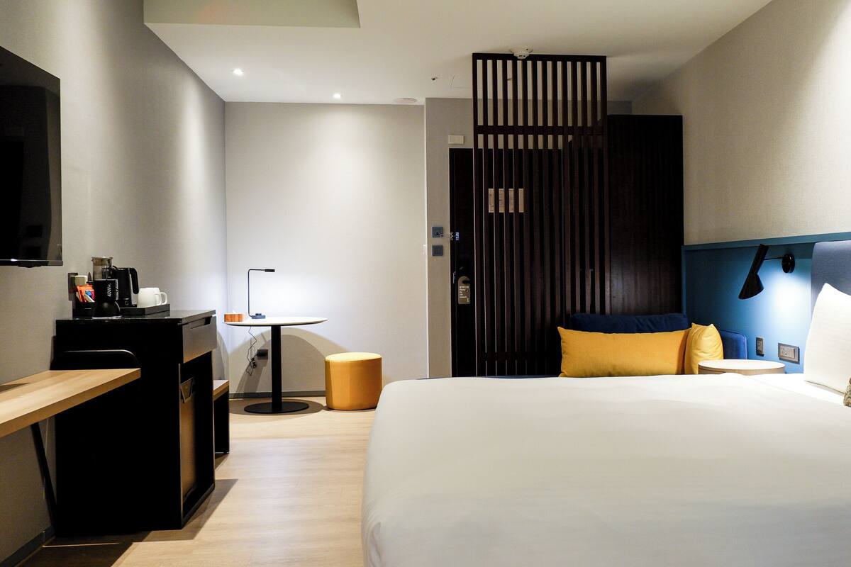 Habitación de hotel con cama, escritorio con cafetera, lámpara, pequeña mesa y pared divisoria.