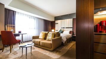 A hotel room with a sofa, a small round table, a red armchair, a bed with a headboard, and a large window with curtains.