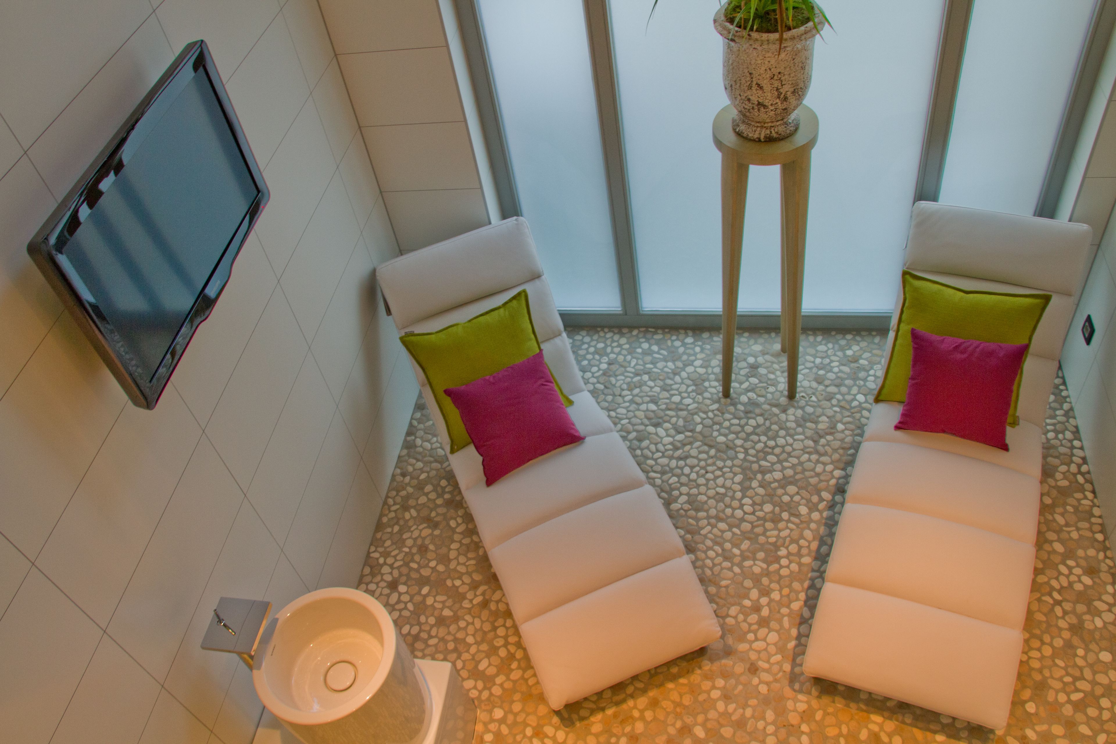 Una sala de spa con dos sillones reclinables, una mesita con una planta, un televisor en la pared y un suelo de baldosas.