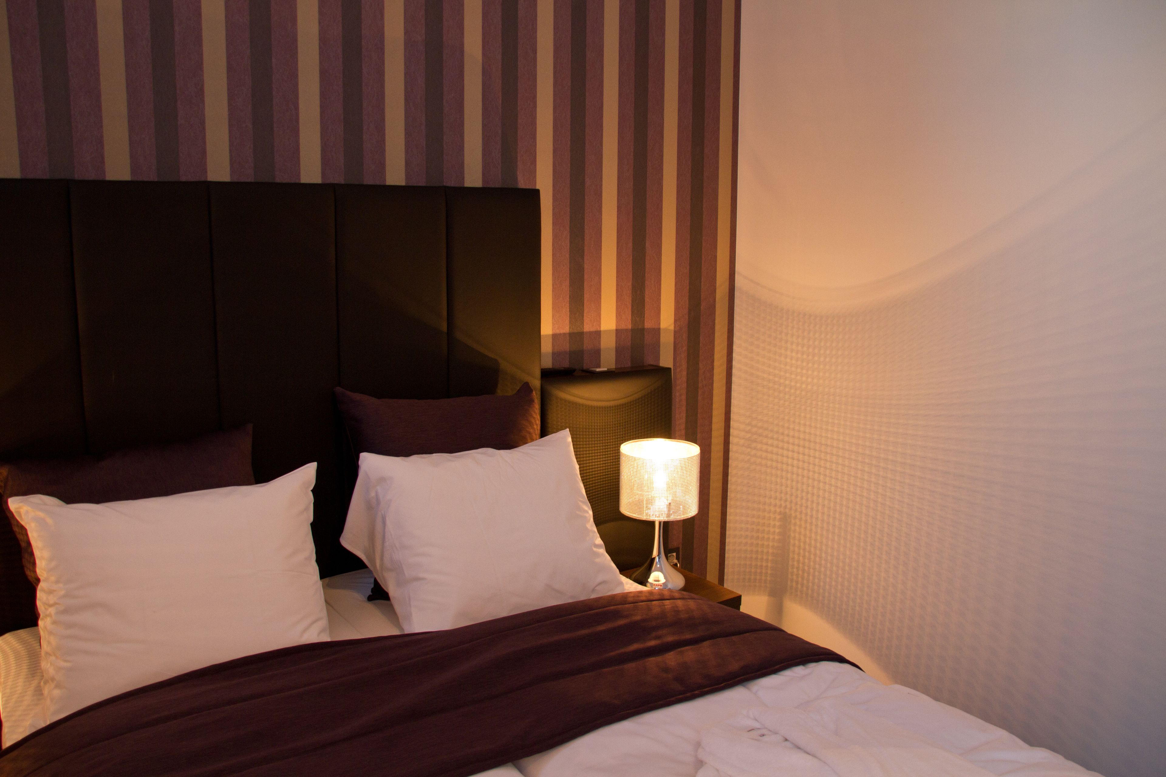 Una cama bien tendida con ropa de cama blanca y marrón, un cabecero con rayas verticales y una lámpara de noche.