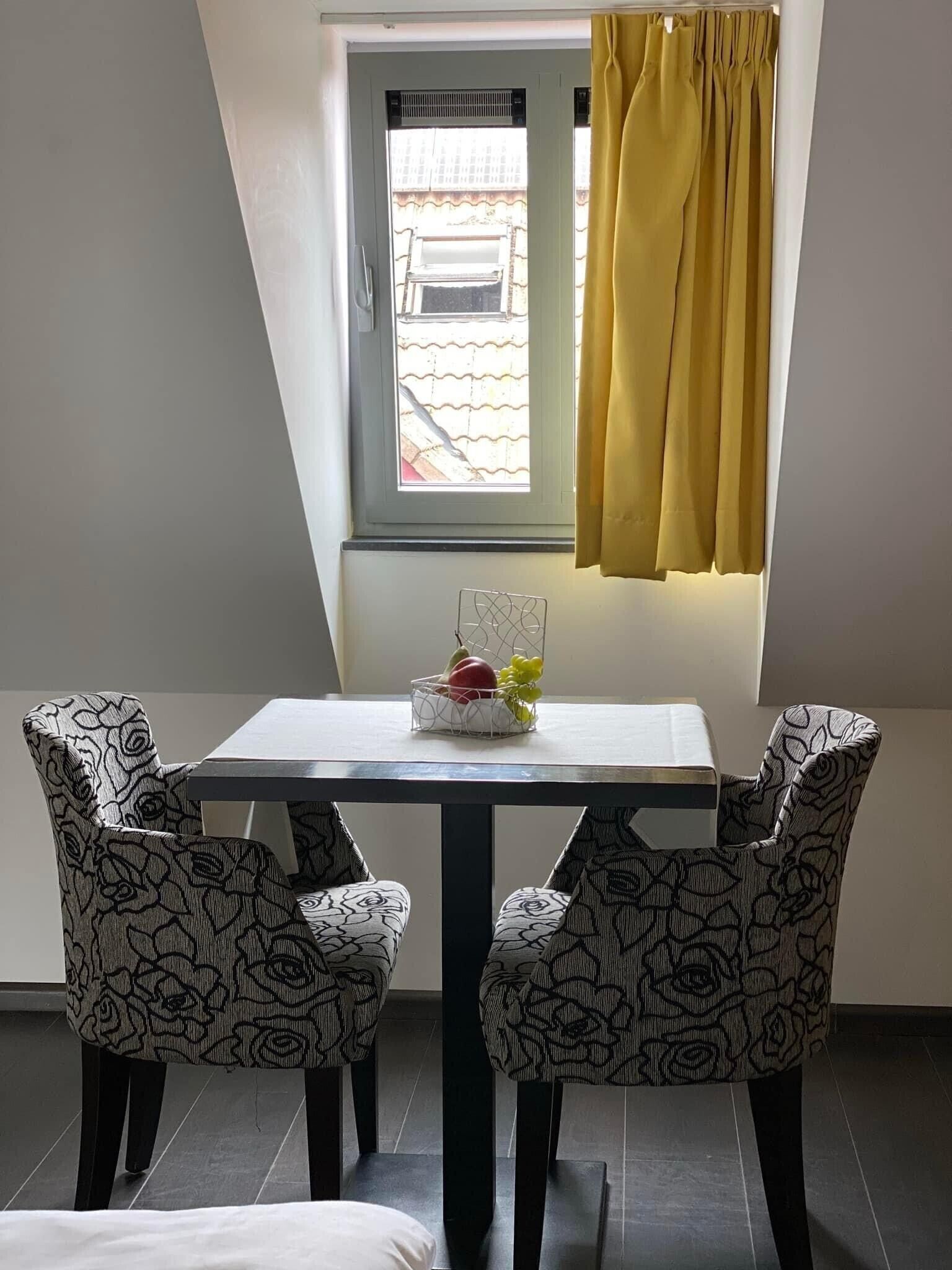 A dining area with a table and four patterned chairs, a window with a yellow curtain, and a fruit basket on the table.