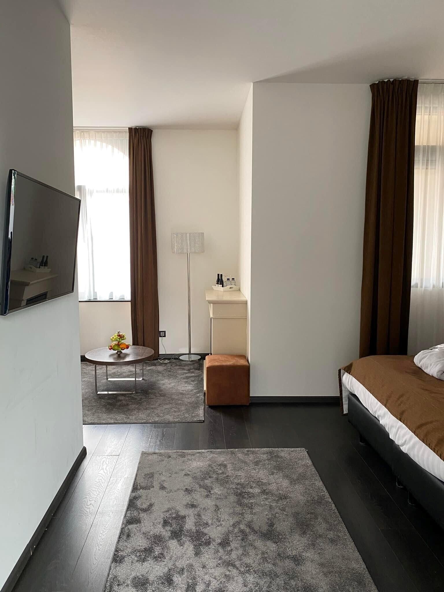 Habitació d'hotel moderna amb un llit, una taula petita amb un ram, una làmpada de peu i un televisor fixat a la paret.