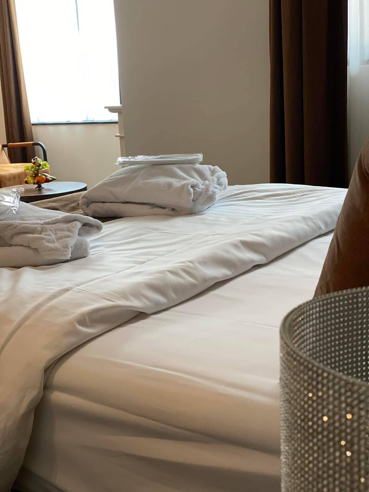 A neatly made bed with white linens and pillows, a bedside table with a fruit bowl, and a window with curtains.