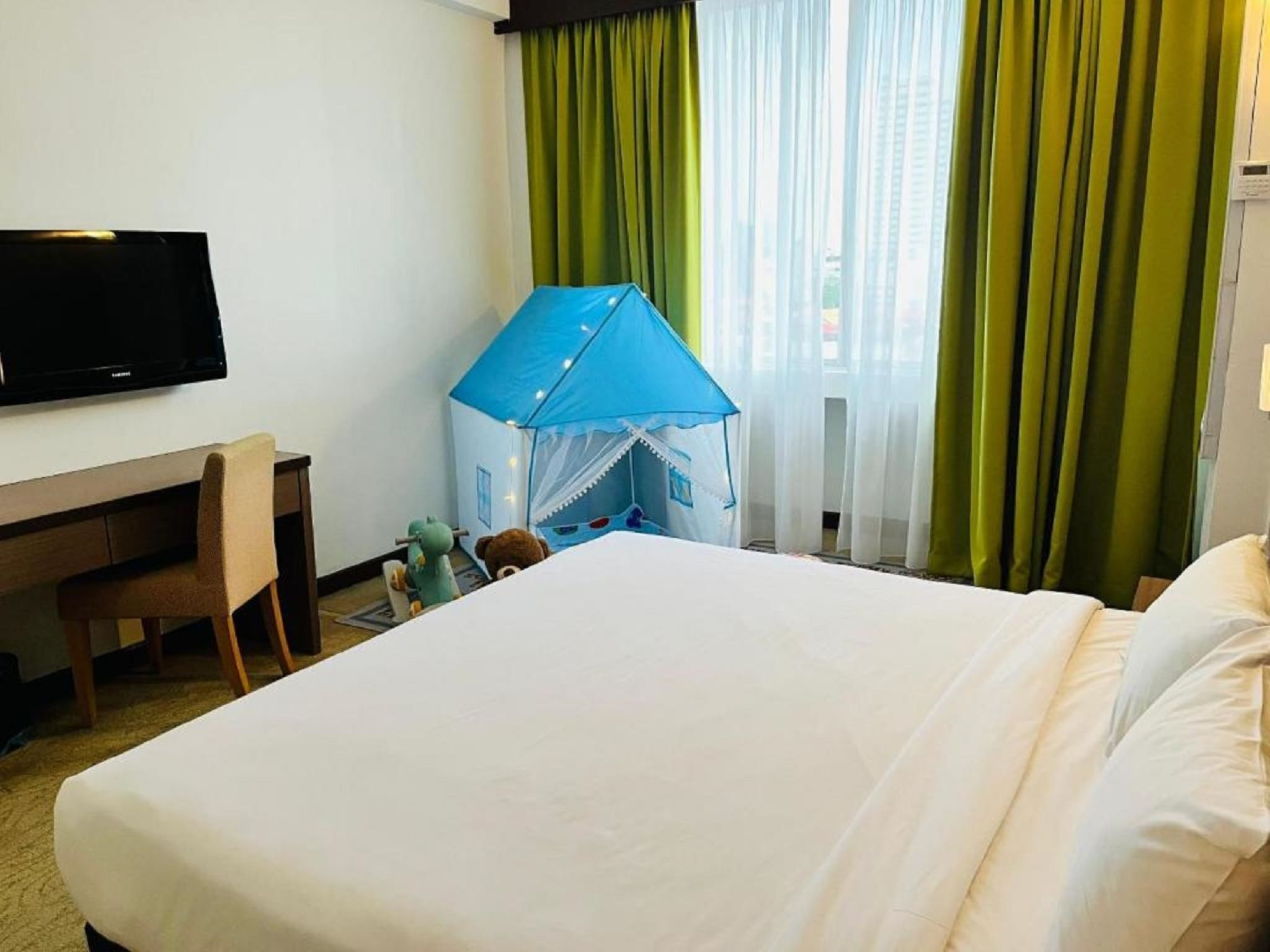 A hotel room with a bed, a desk, a television, a tent-like structure, and green curtains.