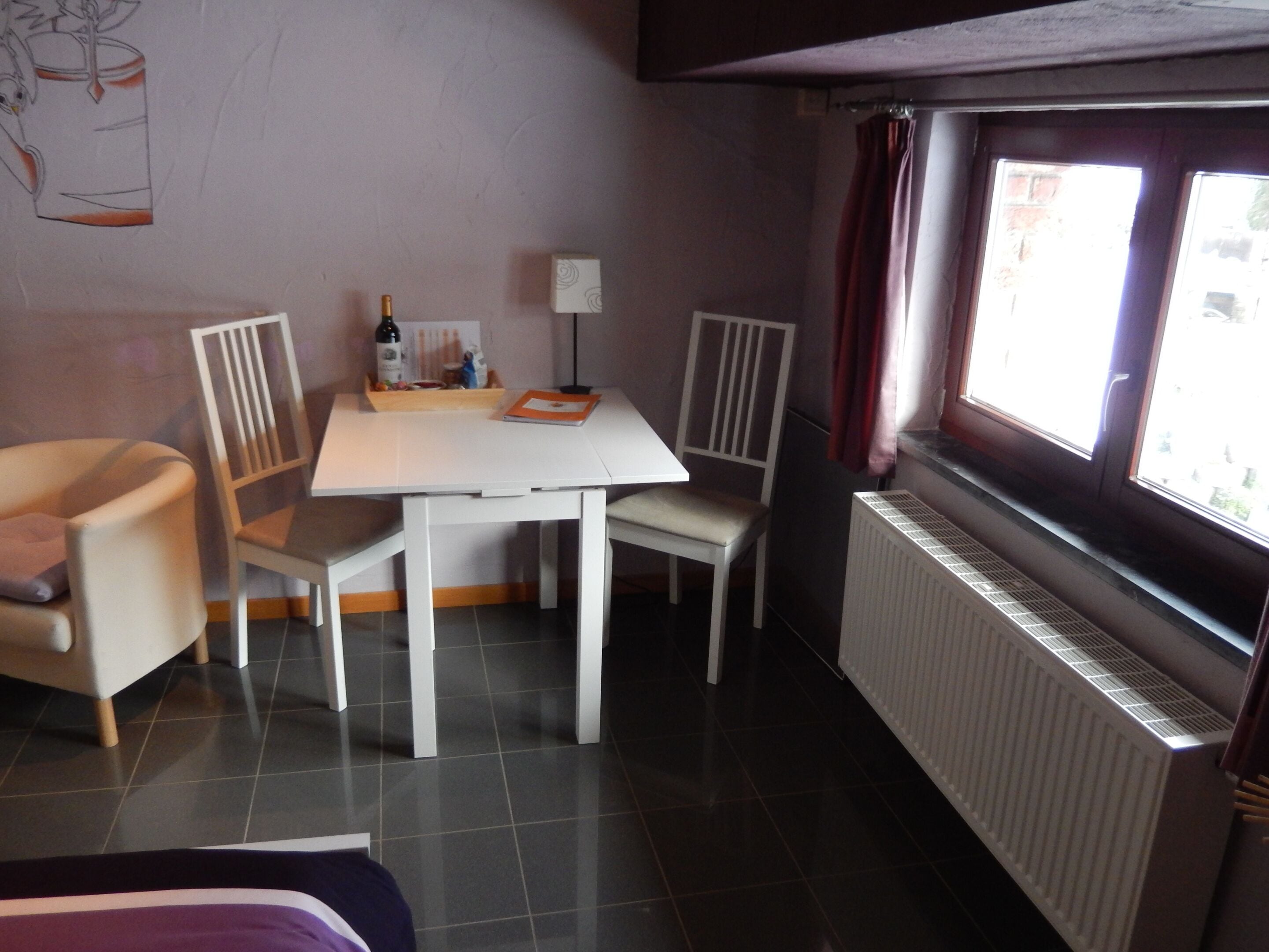 A small, well-lit room with a white table, two chairs, a radiator, and a window with curtains.