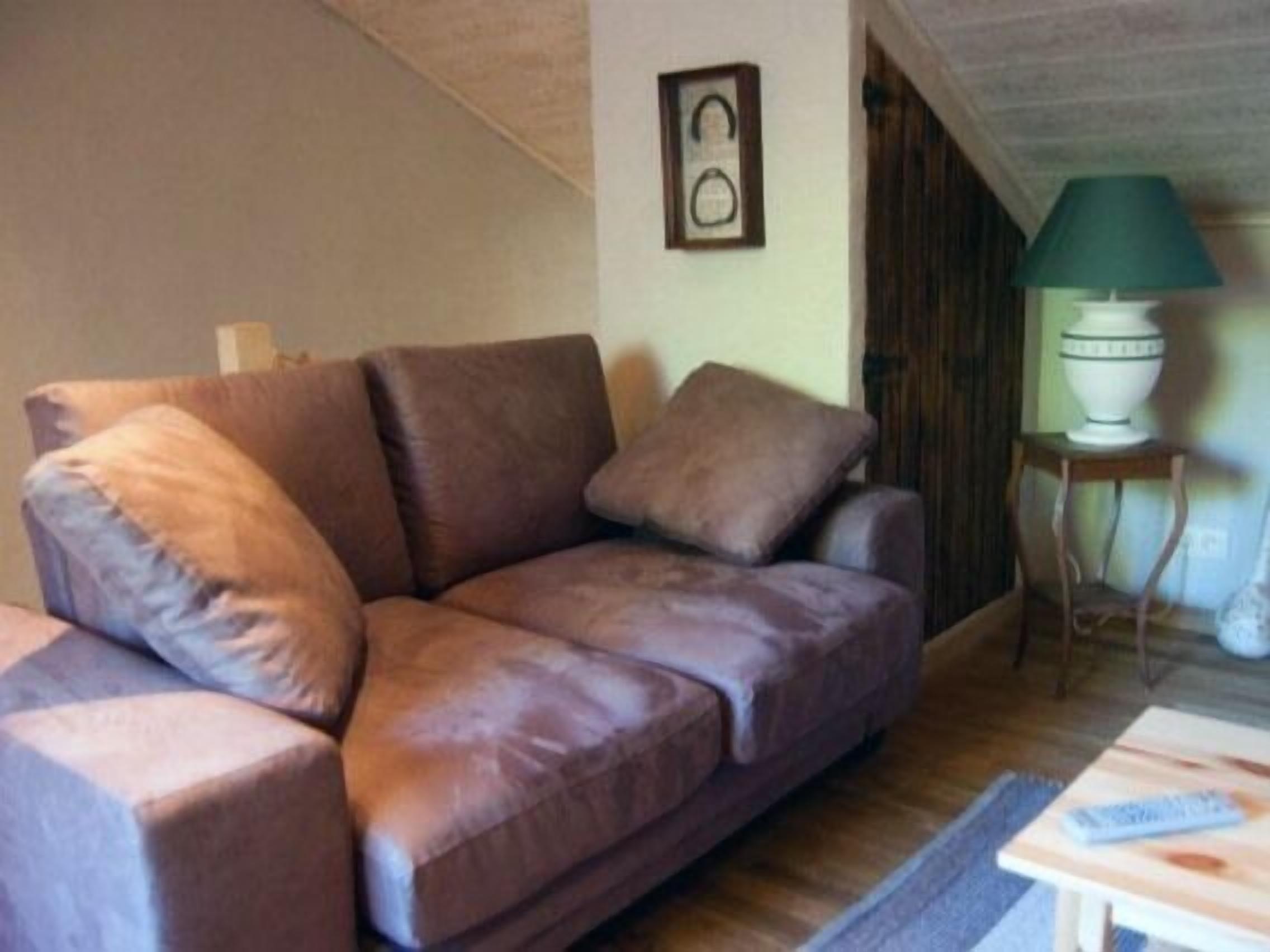 A brown leather sofa with cushions, a small table with a green lamp, and a framed picture on the wall.