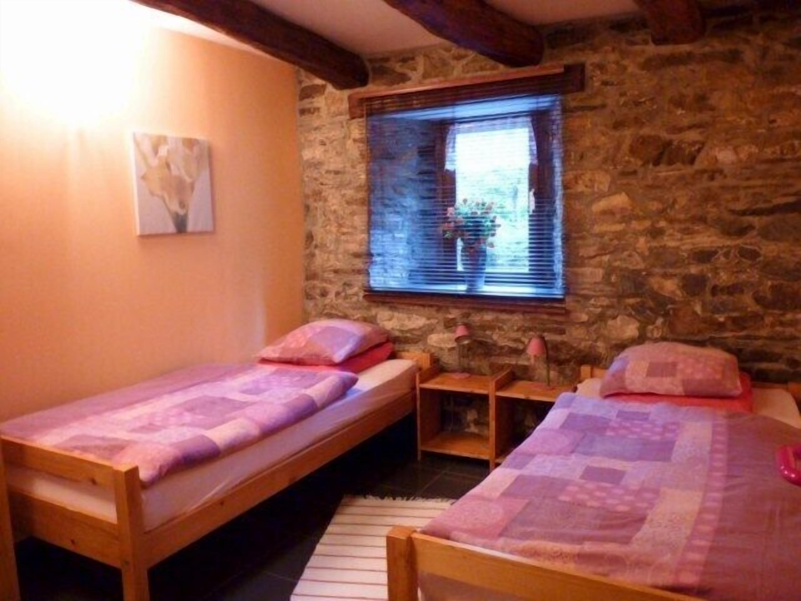 Une chambre avec deux lits en bois, un mur en pierre, une fenêtre avec des stores et une petite table avec une lampe.