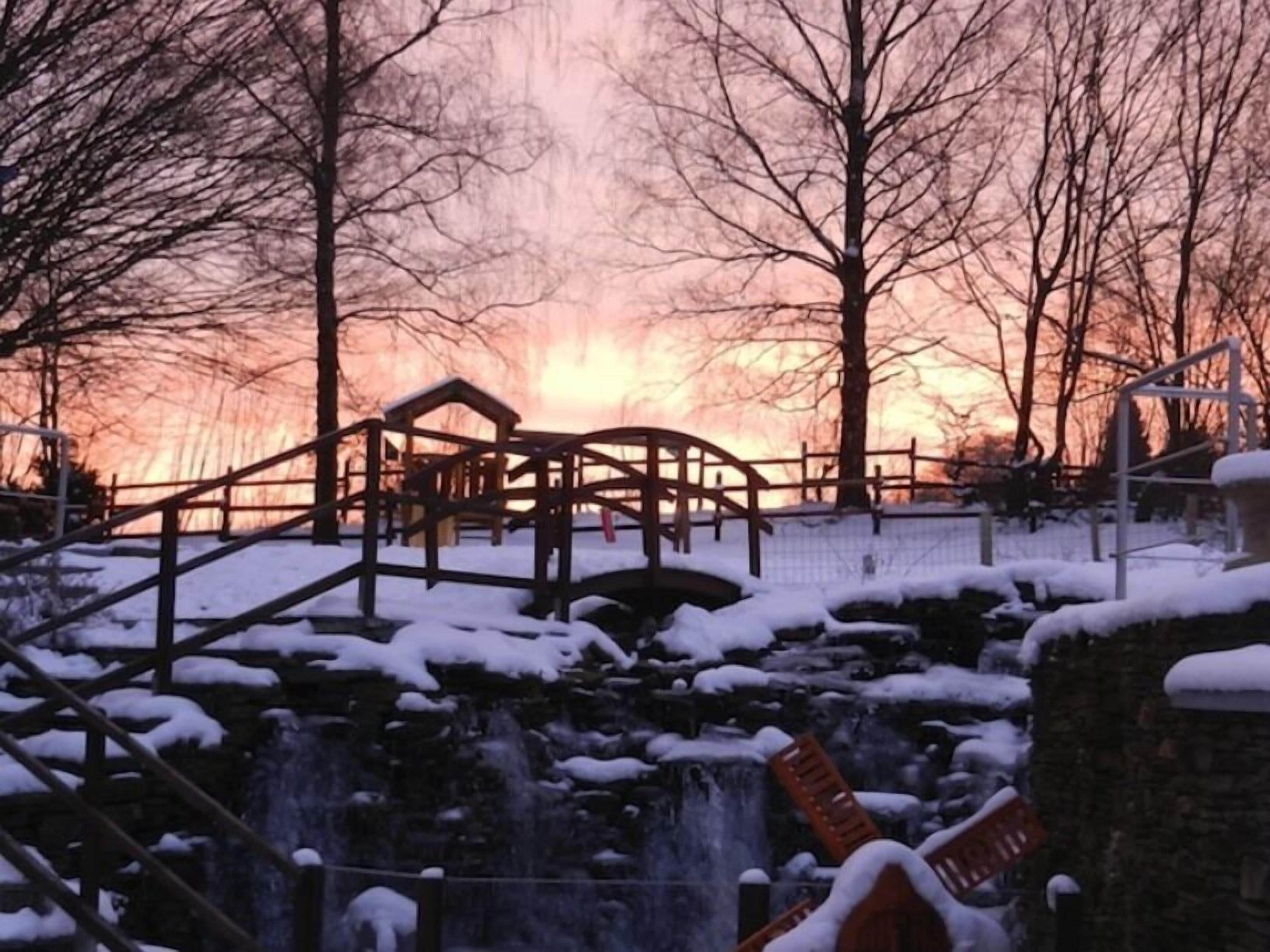 Ein Park mit einer Holzbrücke, einem kleinen Wasserfall und schneebedeckten Bänken.