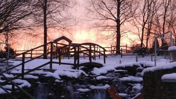 Ein Park mit einer Holzbrücke, einem kleinen Wasserfall und schneebedeckten Bänken.