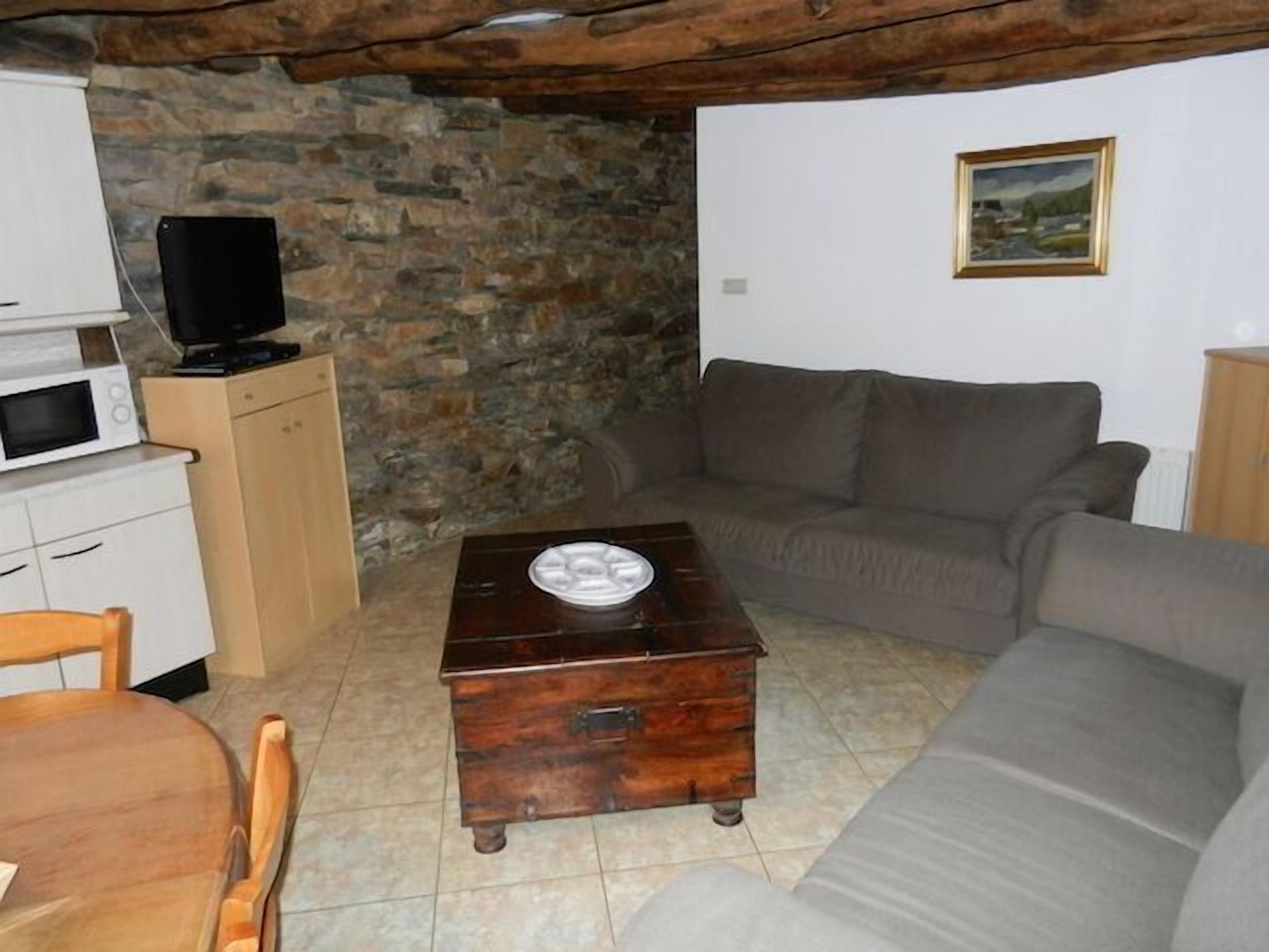 A cozy living room with a stone wall, a wooden chest, a gray sofa, and a television on a cabinet.
