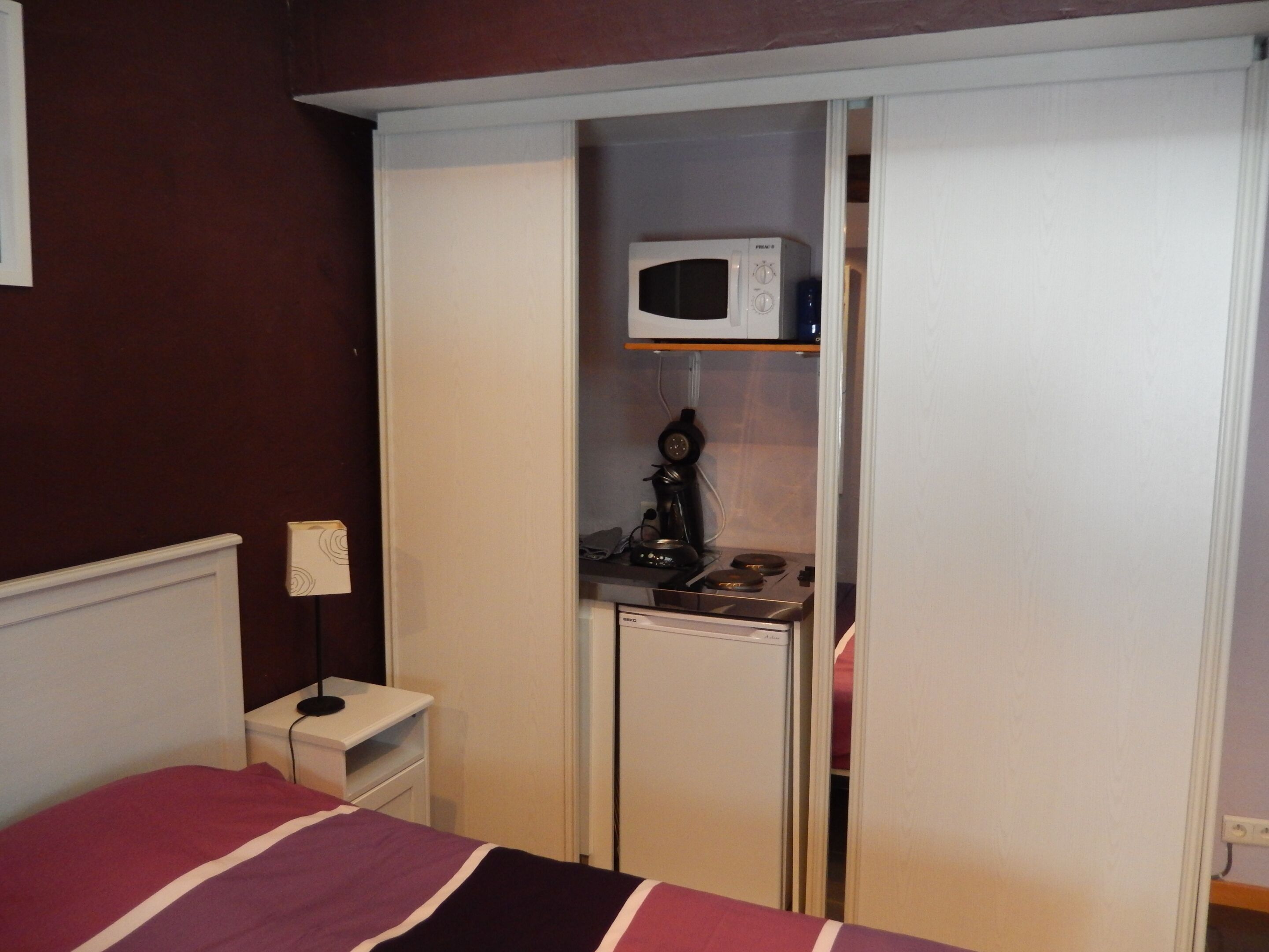 Un dormitorio con cama, mesita de noche, armario, microondas y cafetera.