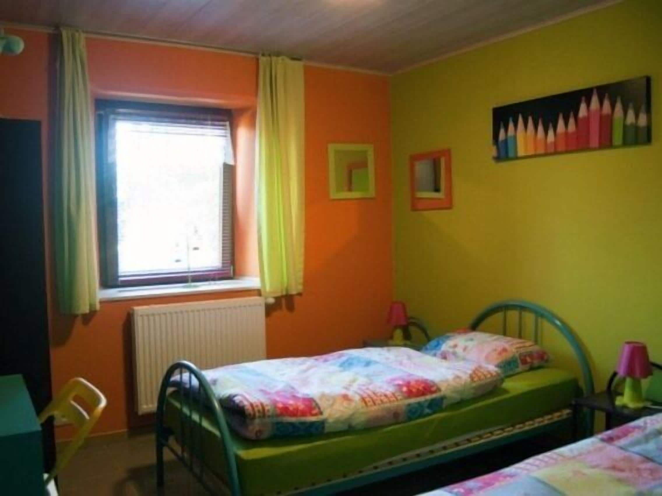 A small, brightly colored room with a single bed, a window with curtains, a radiator, and a wall with a colorful painting.