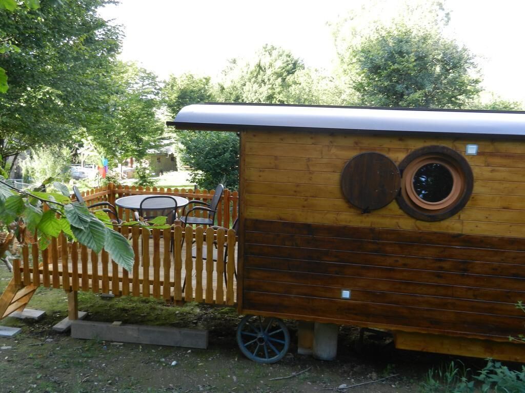 Une nichée à oiseaux en bois, dotée d’un toit incliné et de deux fenêtres rondes, située dans un jardin avec une clôture en bois et une table entourée de chaises.