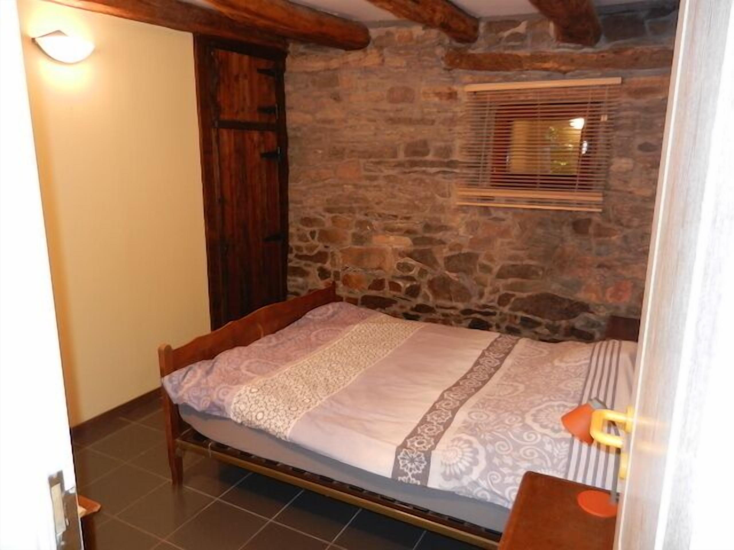 Une petite pièce accueillante avec un mur en pierre, une porte en bois, une fenêtre avec des stores et un lit recouvert d’une literie à motifs.