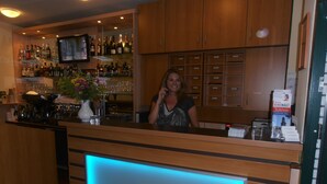 A bar area with wooden cabinets, a TV mounted on the wall, and a blue-lit reception desk. - ALTAN Hotel (Hamburg)
