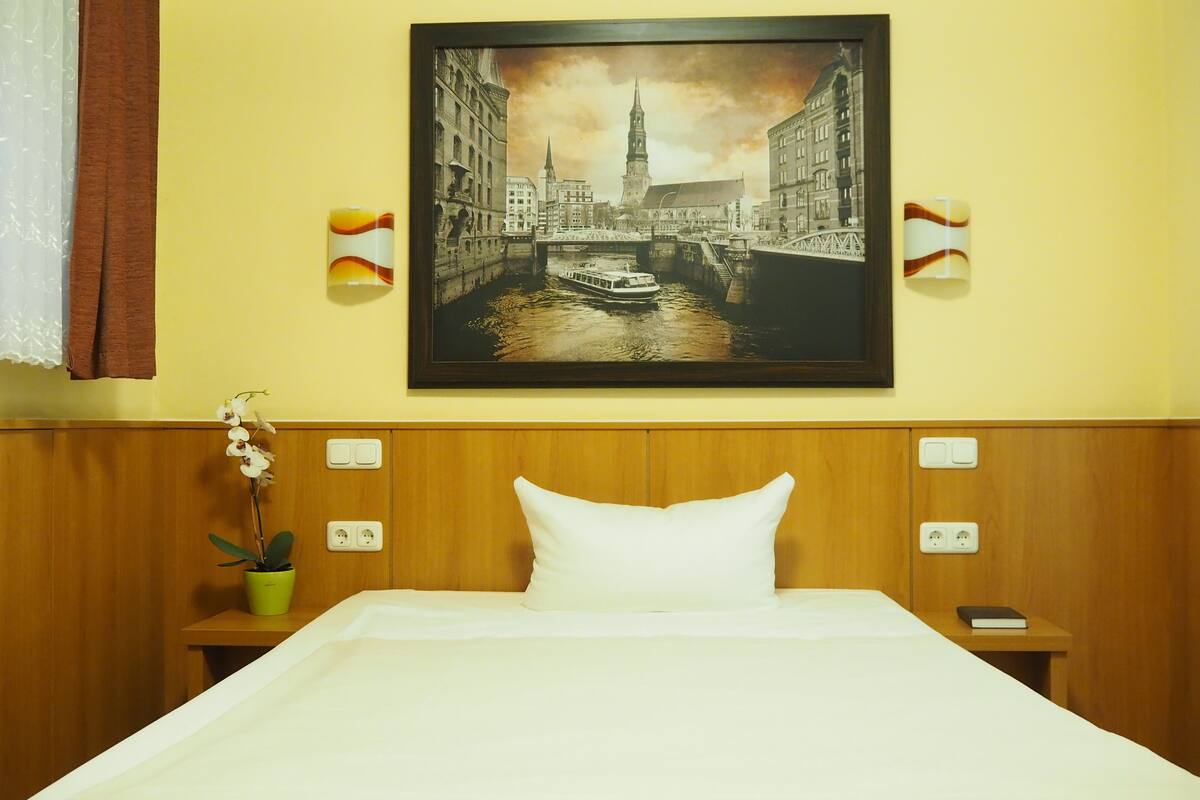 Una camera d'hotel con un letto, un comodino con una lampada, un quadro incorniciato che raffigura un panorama urbano e un'orchidea in vaso.