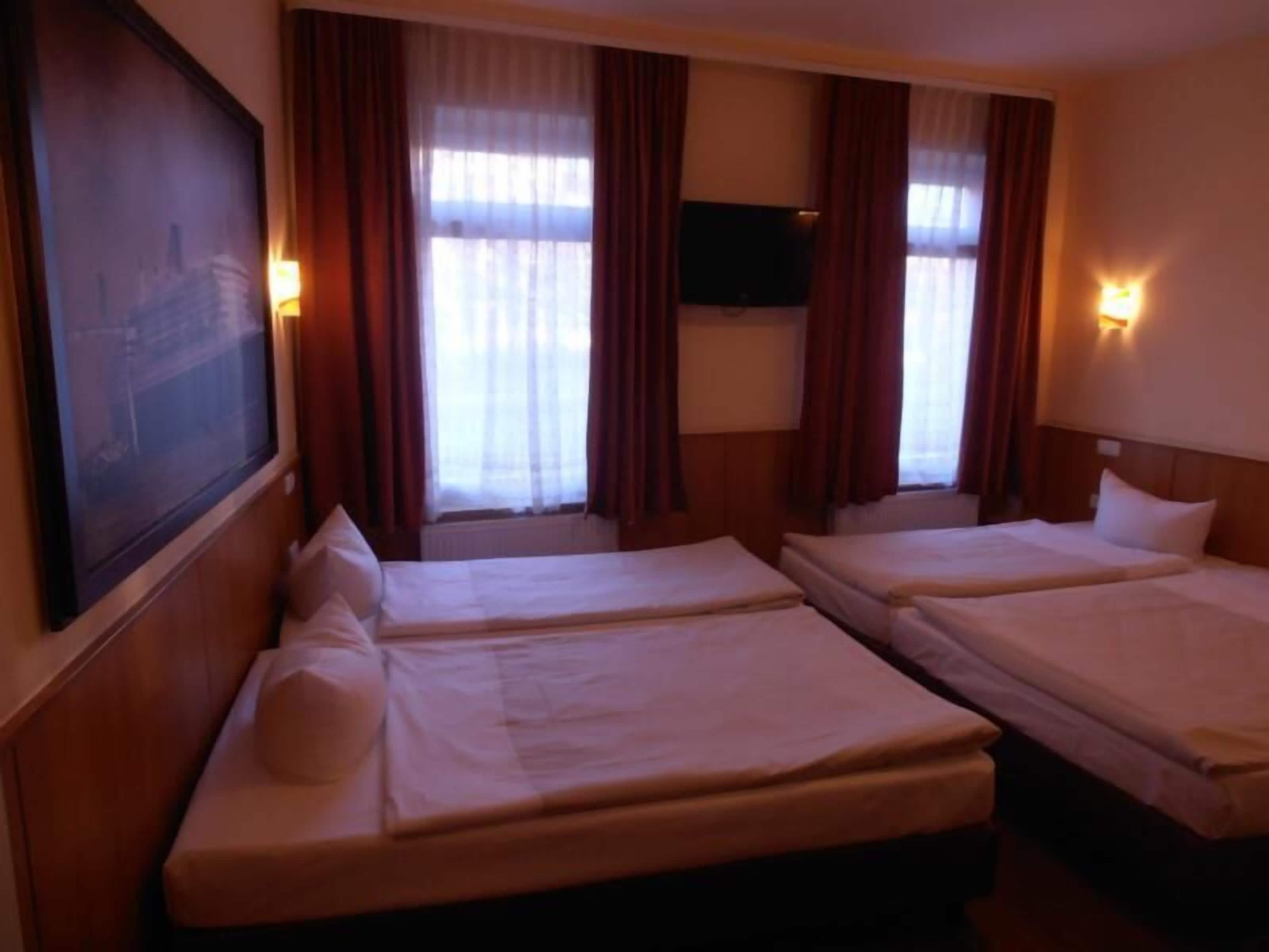 A hotel room with two single beds, a large window with curtains, a flat-screen TV mounted on the wall, and a painting on the wall.