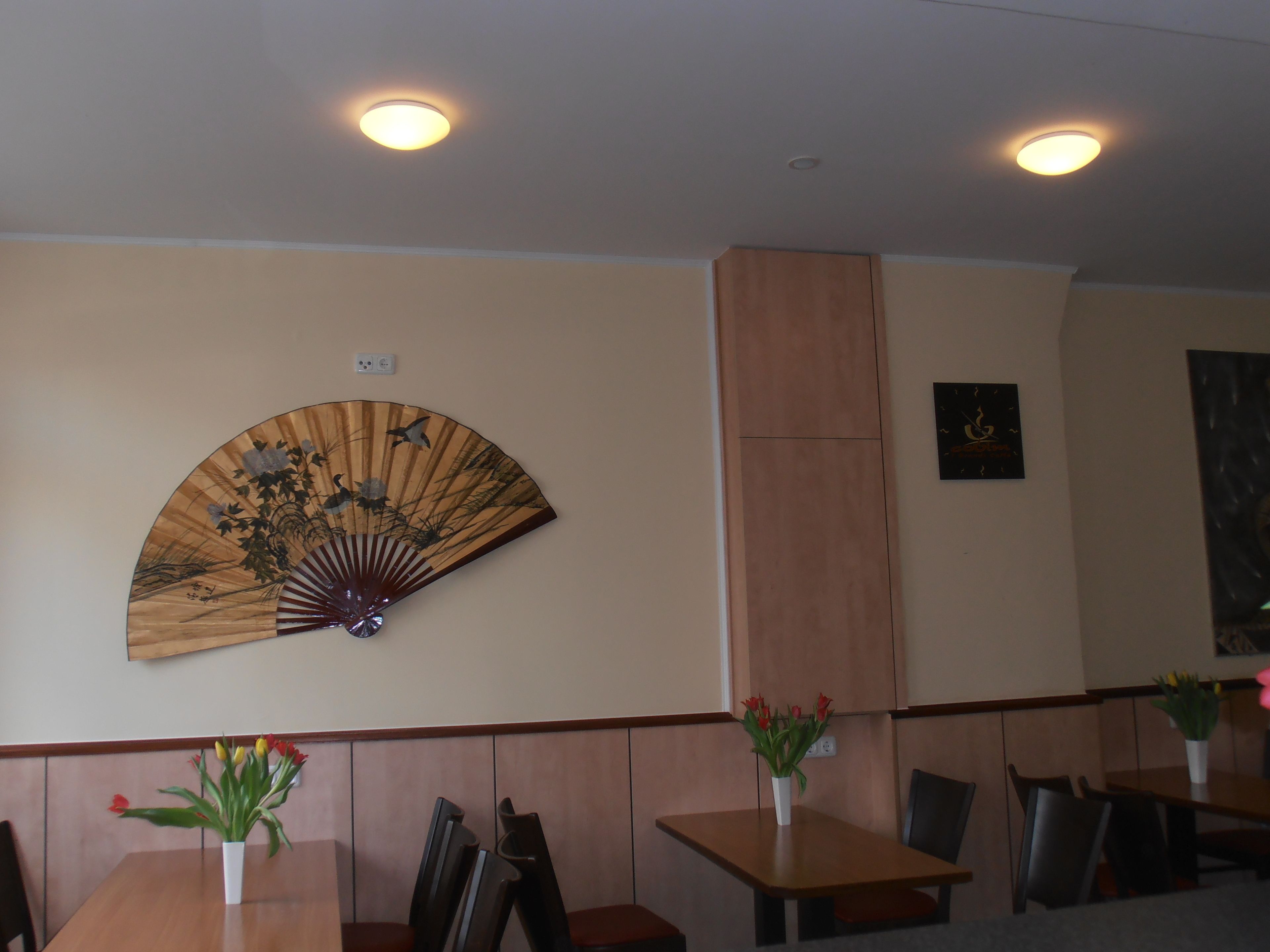 Un comedor con mesas y sillas de madera, un ventilador de pared y un adorno colgante en la pared.
