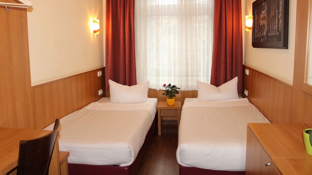 A hotel room with two single beds, wooden headboards, a small table with a plant, and a framed picture on the wall. - ALTAN Hotel (Hamburg)