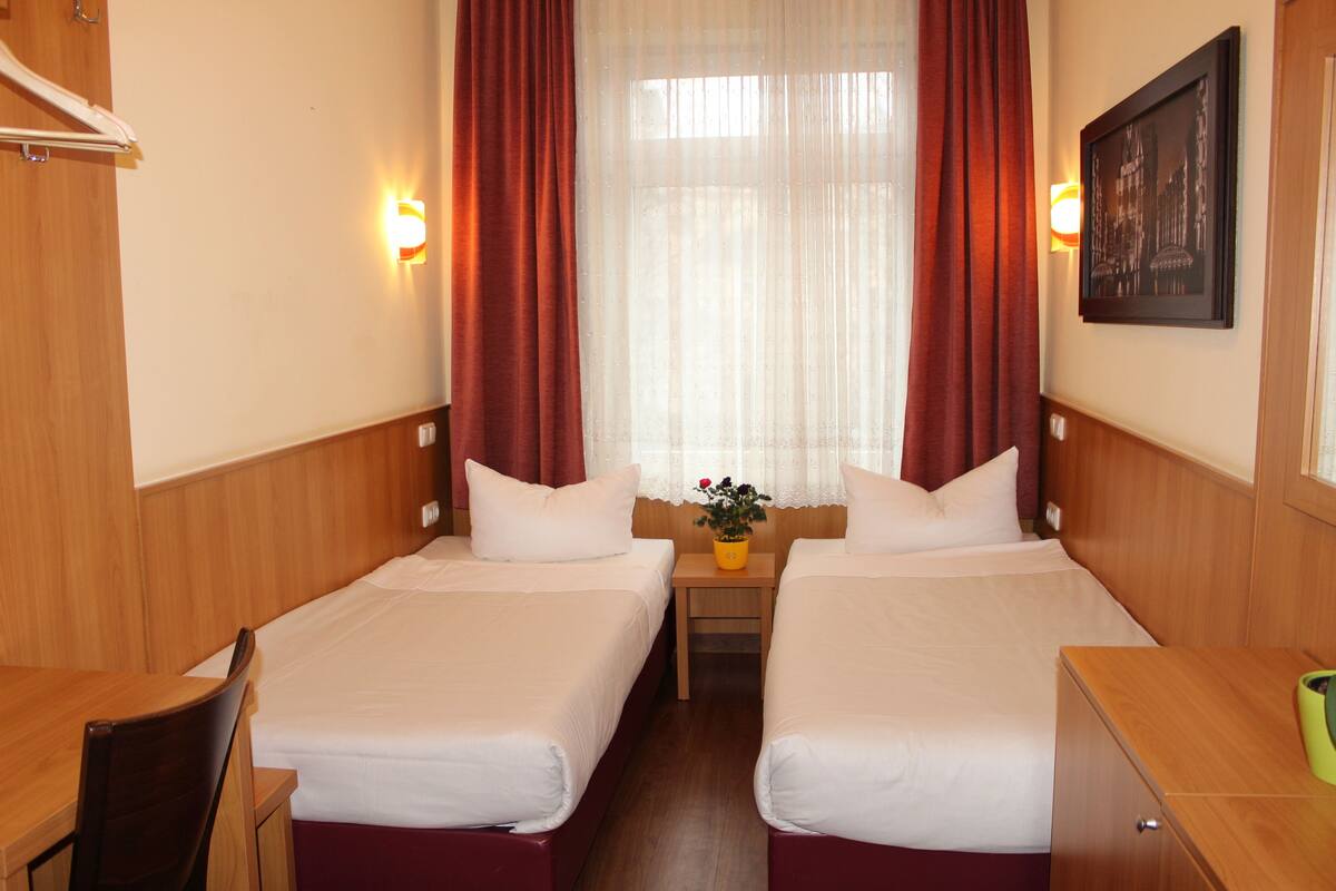 Camera d'albergo con due letti singoli, testiere in legno, un tavolino con una pianta e un quadro appeso al muro.