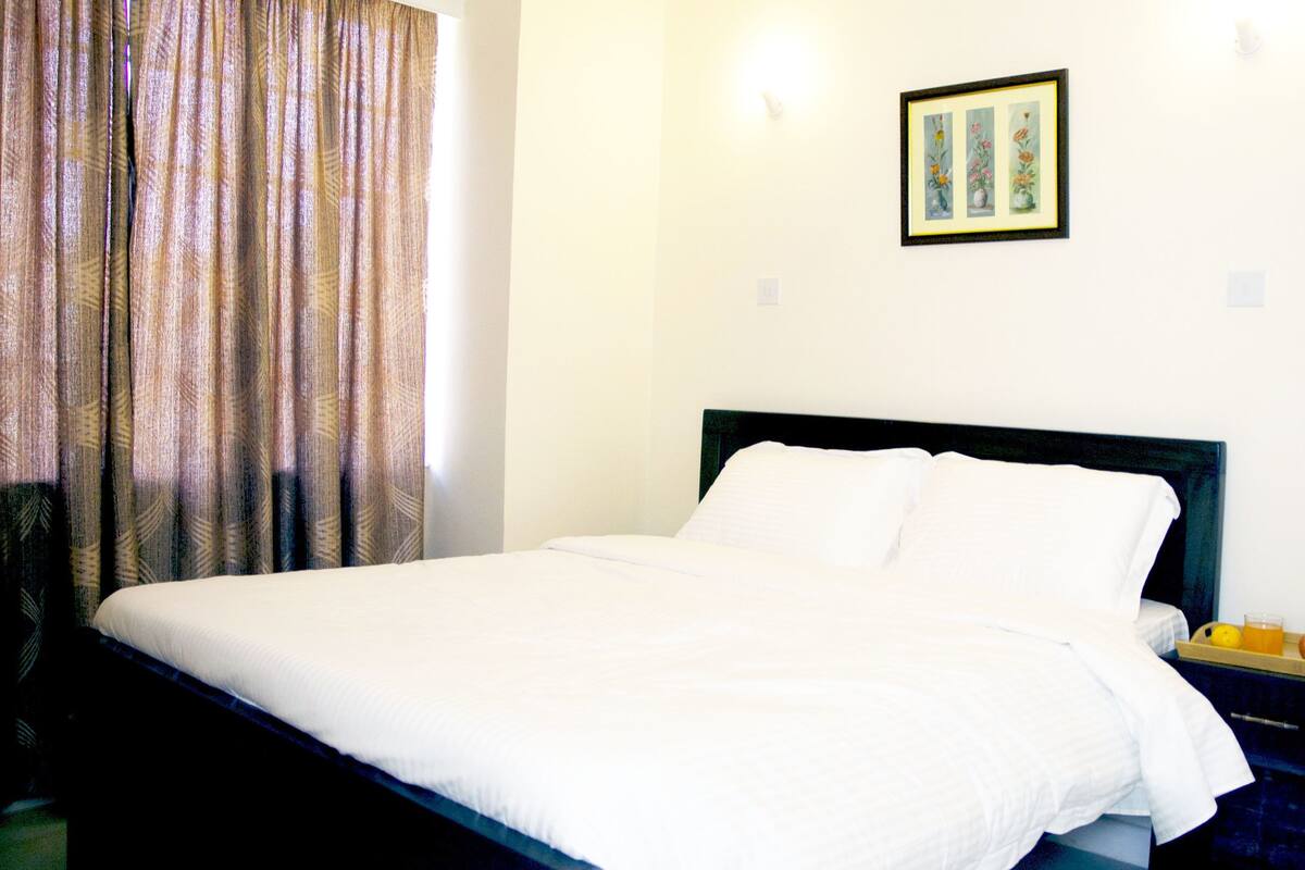 A neatly made bed in a room with a patterned curtain, a framed picture on the wall, and a tray of fruit on the nightstand.